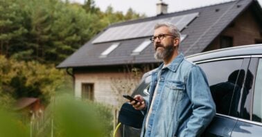barbat langa o masina incarcata electric cu o casa cu panouri solare in fundal