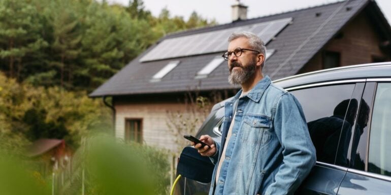 barbat langa o masina incarcata electric cu o casa cu panouri solare in fundal